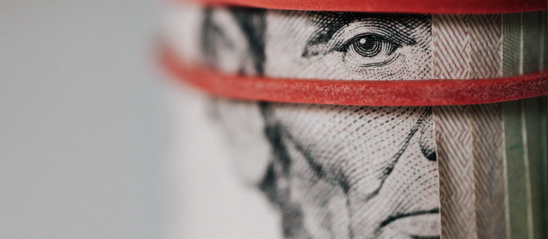 Closeup View Of A Roll Of Five-dollar Bills Held Together By A Red Rubber Band That Frames Abraham Lincoln's Eyes, As If He Is Watching The Viewer.